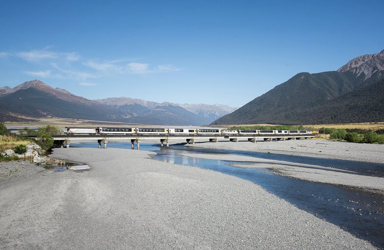 TranzAlpine train crossing a summery Waimakariri River amidst the mountains of Arthur's Pass National Park.